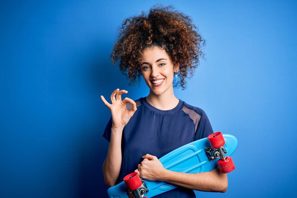 Молодая спортивная женщина с вьющимися волосами и пирсинг держа коньки на синем фоне делает ОК знак с пальцами, отличный символ
 - Фото, изображение