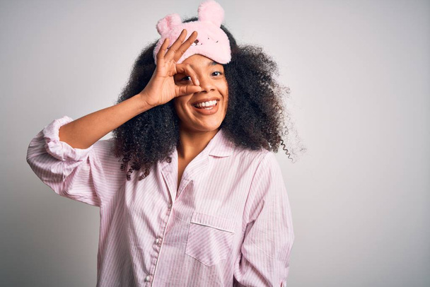 Joven mujer afroamericana con cabello afro usando máscara para dormir y pijama rosa haciendo un buen gesto con la mano sonriente, ojos mirando a través de los dedos con la cara feliz
. - Foto, Imagen