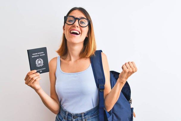 Beautiful redhead student woman wearing backpack and holding passport of italy screaming proud and celebrating victory and success very excited, cheering emotion - 写真・画像