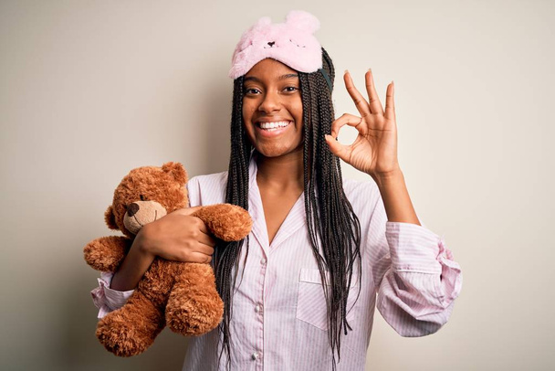 Nuori afrikkalainen amerikkalainen nainen yllään pyjama ja silmä naamio halaus nallekarhu tekee ok merkki sormilla, erinomainen symboli
 - Valokuva, kuva