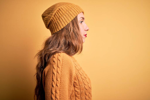 Молодая красивая блондинка носит случайный свитер и шерстяную шапку на желтом фоне глядя в сторону, расслабиться профиль позировать с естественным лицом с уверенной улыбкой
. - Фото, изображение