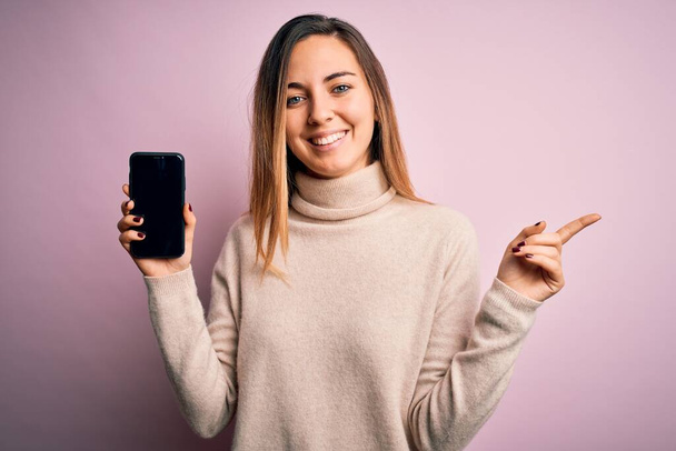 Молодая красивая брюнетка женщина, держащая смартфон показывая экран на розовом фоне очень счастлив указывая рукой и пальцем в сторону
 - Фото, изображение