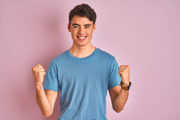 Tiener jongen draagt casual t-shirt staan over blauw geïsoleerde achtergrond vieren verrast en verbaasd voor succes met opgeheven armen en open ogen. Winnaars concept. - Foto, afbeelding