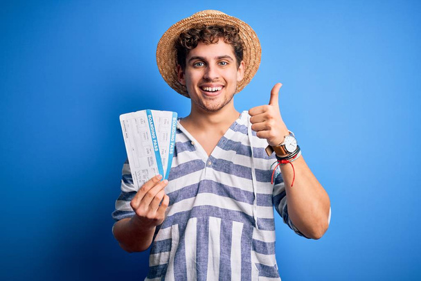 Young blond tourist man with curly hair on vacation wearing summer hat holding boarding pass happy with big smile doing ok sign, thumb up with fingers, excellent sign - Foto, Imagem