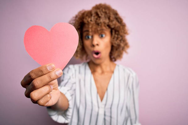Young african american romantic woman with curly hair holding paper heart shape scared in shock with a surprise face, afraid and excited with fear expression - Foto, Bild