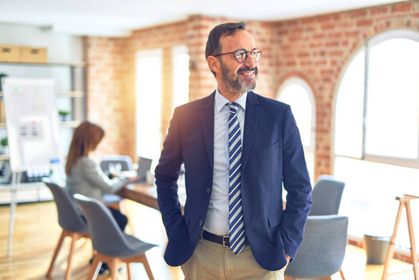 Middle age handsome businessman wearing glasses   standing at the office looking away to side with smile on face, natural expression. Laughing confident. - Photo, Image