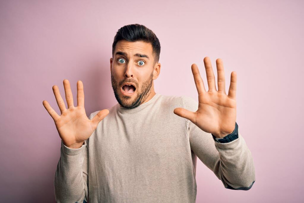 Young handsome man wearing casual sweater standing over isolated pink background afraid and terrified with fear expression stop gesture with hands, shouting in shock. Panic concept. - Photo, Image