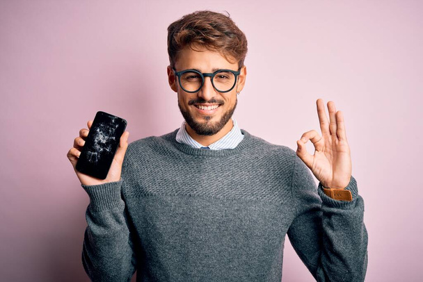 Mladý muž s vousy nosí brýle drží zlomený a prasklý smartphone dělá v pořádku znamení s prsty, vynikající symbol - Fotografie, Obrázek