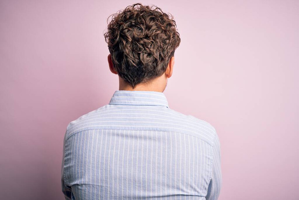 Young blond handsome man with curly hair wearing striped shirt over white background standing backwards looking away with crossed arms - Photo, Image