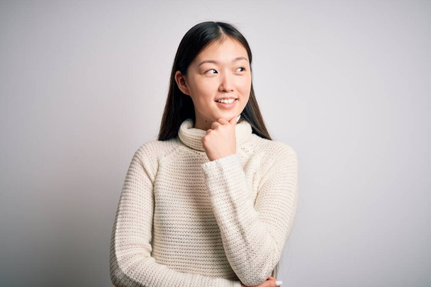 Jonge mooie Aziatische vrouw draagt casual trui staan over geïsoleerde achtergrond met de hand op kin denken over vraag, pensive expressie. Lachend met een bedachtzaam gezicht. Twijfelachtig concept. - Foto, afbeelding