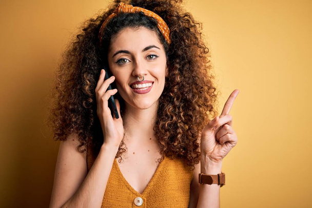 Kıvırcık saçlı ve piercingli genç bir kadın akıllı telefondan konuşuyor. Bir fikir ya da soru işareti ile şaşırdı. - Fotoğraf, Görsel
