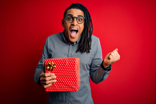 赤の孤立した背景の上にプレゼントボックスを保持しているアフリカ系アメリカ人の男は誇りを叫び、勝利と成功を祝う非常に興奮し、応援感情 - 写真・画像