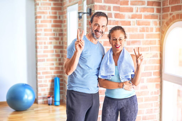 Couple sportif d'âge moyen debout avant de faire de l'exercice à la salle de gym souriant avec un clin d'oeil heureux visage à la caméra faisant signe de victoire. Numéro deux
. - Photo, image