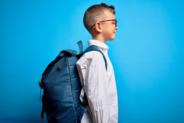 Młody mały biały uczeń dzieciak nosi inteligentne okulary i torbę szkolną na niebieskim tle patrząc na bok, relaks poza profil z naturalną twarzą z pewnym uśmiechem. - Zdjęcie, obraz
