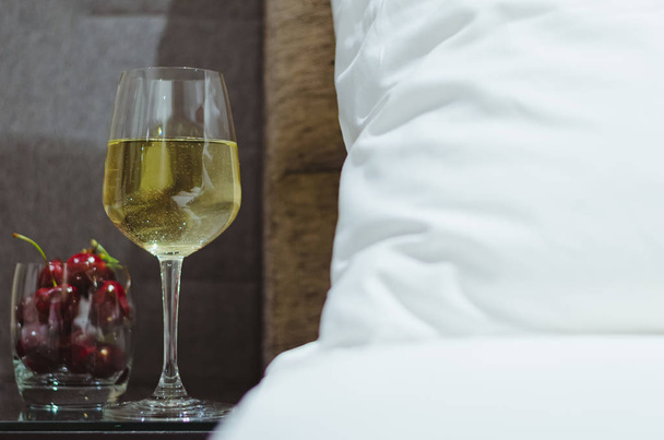 A glass of white wine with cherry fruits put on table besides bed with white bedsheet. - Fotografie, Obrázek