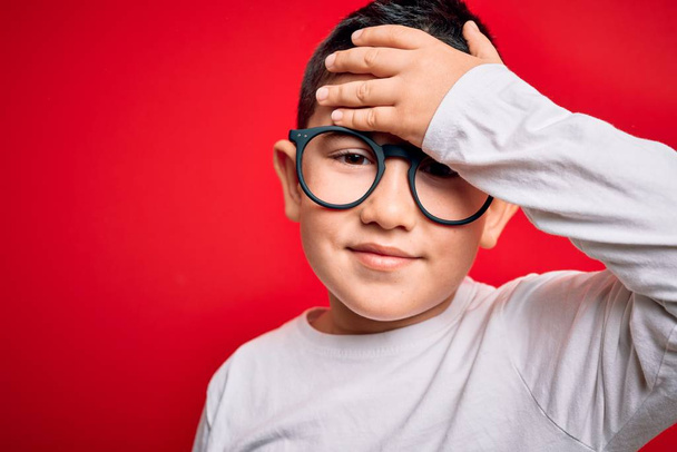 Young little smart boy kid wearing nerd glasses over red isolated background stressed with hand on head, shocked with shame and surprise face, angry and frustrated. Fear and upset for mistake. - Photo, Image