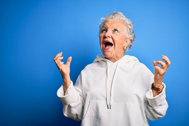 Senior gyönyörű sportos nő fehér pulóvert visel elszigetelt kék háttér őrült és őrült kiabálás és kiabálás agresszív kifejezés és karok emelt. Frusztrációs koncepció. - Fotó, kép