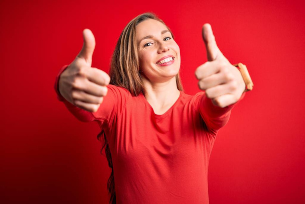 手で積極的なジェスチャーを行うことを承認孤立した赤い背景の上に立ってカジュアルなTシャツを着て若い美しいブロンドの女性,成功のために笑顔と幸せを親指アップ.勝者のジェスチャー. - 写真・画像