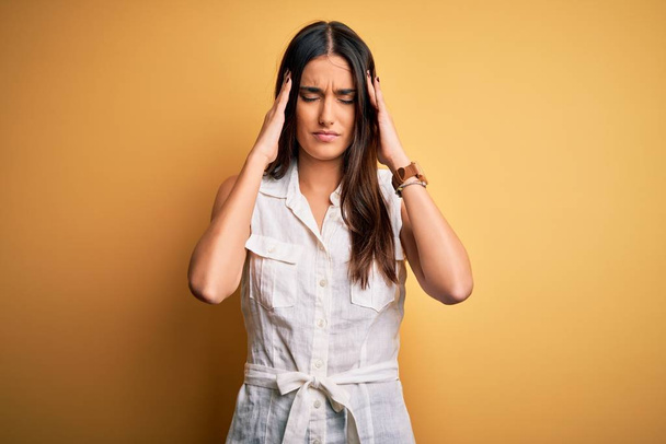 Young beautiful brunette woman on vacation wearing casual dress over yellow background suffering from headache desperate and stressed because pain and migraine. Hands on head. - Photo, image