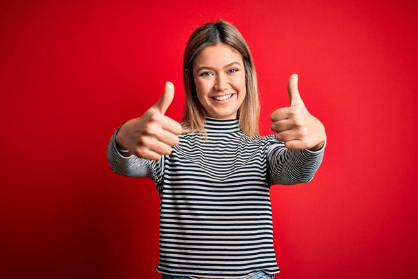 若い美しいブロンドの女性は、赤い隔離された背景にカジュアルなストライプセーターを着て、手で積極的なジェスチャーを行うことを承認し、親指を上げて笑顔と成功のために幸せ。勝者のジェスチャー. - 写真・画像