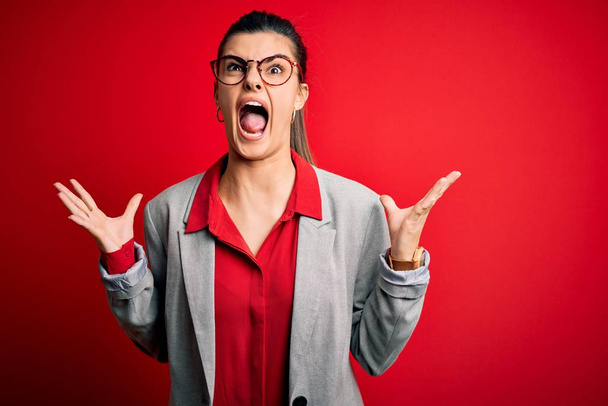 Young beautiful brunette businesswoman wearing jacket and glasses over red background crazy and mad shouting and yelling with aggressive expression and arms raised. Frustration concept. - Φωτογραφία, εικόνα