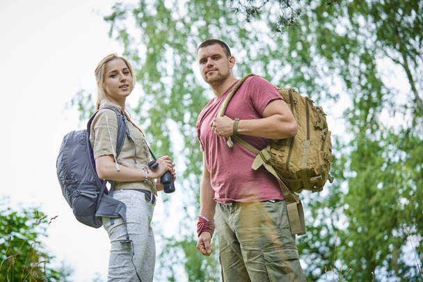 two travelers with binoculars and backpacks resting in forest at sunny day  - Photo, Image