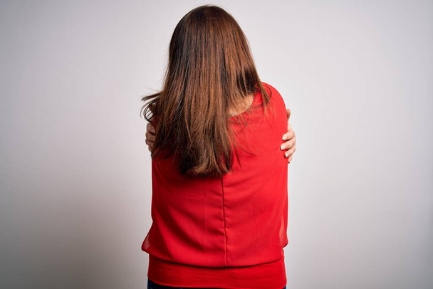 Krásná bruneta plus velikost žena nosí ležérní červené tričko přes izolované bílé pozadí objímající se šťastný a pozitivní od pozpátku. Sebeláska a sebepéče - Fotografie, Obrázek