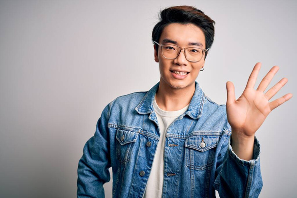 Young handsome chinese man wearing denim jacket and glasses over white background showing and pointing up with fingers number five while smiling confident and happy. - Φωτογραφία, εικόνα