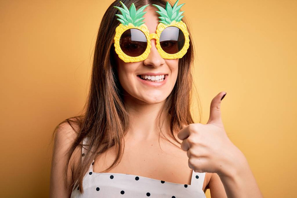 Jeune belle femme brune portant des lunettes de soleil à l'ananas sur fond jaune heureuse avec un grand sourire faisant signe ok, pouce levé avec les doigts, excellent signe
 - Photo, image