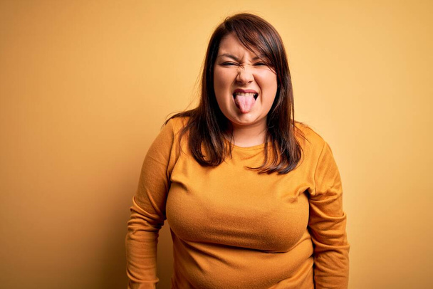 Брюнетка plus size женщина в случайном свитере на изолированном желтом фоне, торчащем языком, довольная смешным выражением лица. Концепция эмоций
. - Фото, изображение