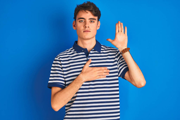 Teenager-Junge in lässigem T-Shirt, der vor blauem Hintergrund steht und mit der Hand auf der Brust und offener Handfläche schwört, einen Treueschwur abzulegen - Foto, Bild