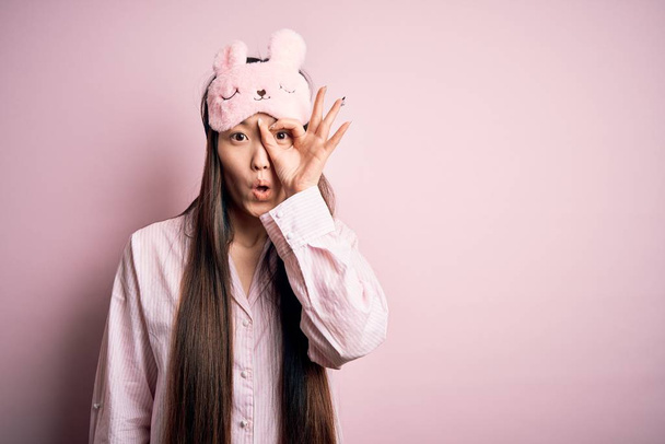 Jeune femme asiatique portant un pyjama et un masque de sommeil sur fond rose isolé faisant un geste correct choqué par le visage surpris, les yeux regardant à travers les doigts. Expression incrédule
. - Photo, image