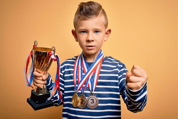 Jonge kleine blanke jongen draagt winnaar medailles en overwinning award trofee over gele achtergrond geïrriteerd en gefrustreerd schreeuwen van woede, gek en schreeuwen met opgeheven hand, woede concept - Foto, afbeelding