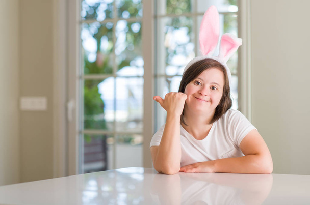 Kobieta z zespołem Downa w domu, nosząca wielkanocne uszy królika wskazujące i pokazujące z kciukiem do góry z uśmiechniętą twarzą. - Zdjęcie, obraz