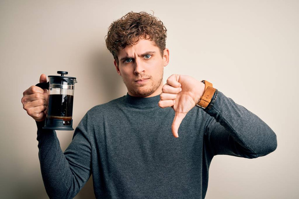 Young blond man with curly hair making coffee using coffemaker over white background with angry face, negative sign showing dislike with thumbs down, rejection concept - Photo, image