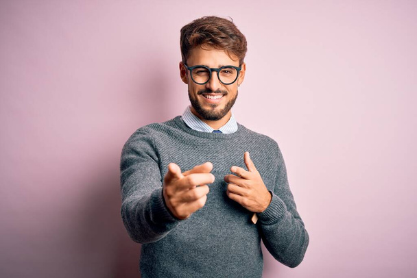 Fiatal jóképű férfi szakállal szemüvegben és pulóverben, rózsaszín háttér felett áll, ujjal mutogatva a kamerára, boldog és vicces arccal. Jó energia és rezgések. - Fotó, kép