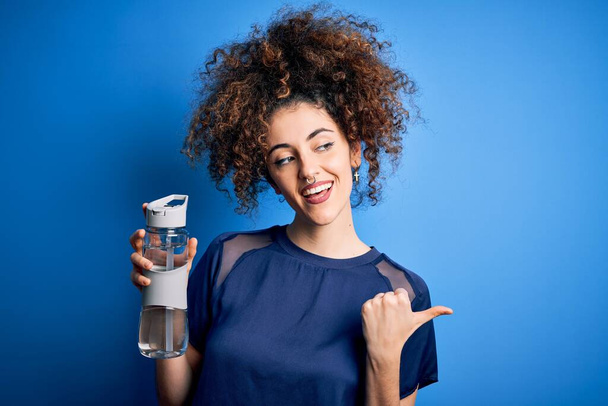 Young beautiful sporty woman with curly hair and piercing doing sport holding bottle of water pointing and showing with thumb up to the side with happy face smiling - Photo, Image
