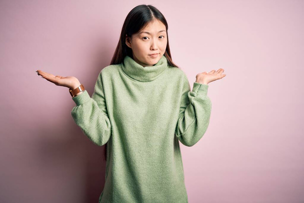 Młoda piękna Azjatka ubrana w zielony zimowy sweter na różowym, osłoniętym tle, nieświadoma i zdezorientowana ekspresja z podniesionymi rękami i rękami. Koncepcja wątpliwości. - Zdjęcie, obraz