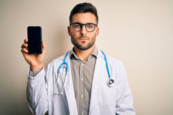 Young doctor man wearing stethoscope showing smartphone screen over isolated background with a confident expression on smart face thinking serious - Fotoğraf, Görsel