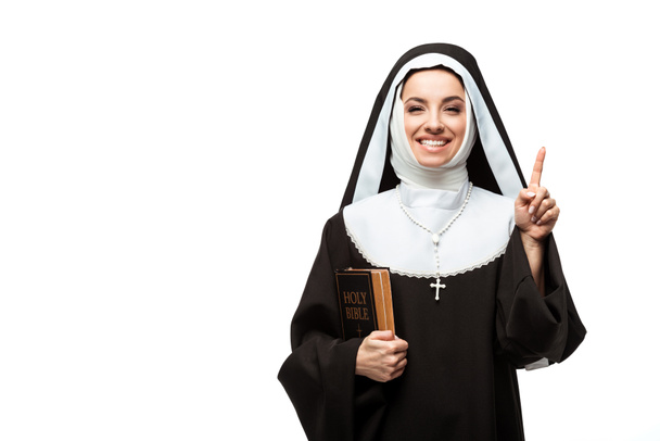 fröhliche Nonne hält Bibel in der Hand und zeigt mit Idee isoliert auf weiß - Foto, Bild