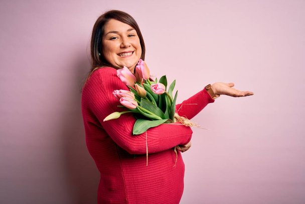 Gyönyörű plusz méret nő gazdaság romantikus csokor természetes tulipán virágok felett rózsaszín háttér mutatva félre a kezét nyitott tenyér mutatja másolás tér, bemutató reklám mosolygó izgatott boldog - Fotó, kép