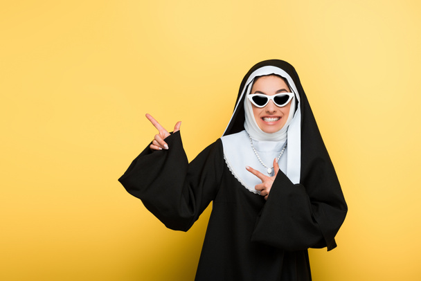 freira alegre atraente em óculos de sol apontando no amarelo
 - Foto, Imagem
