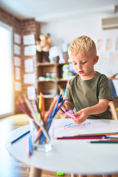 Молодой кавказский ребенок играет в детском саду, рисуя цветными карандашами. Дошкольный мальчик счастлив в игровой комнате
. - Фото, изображение