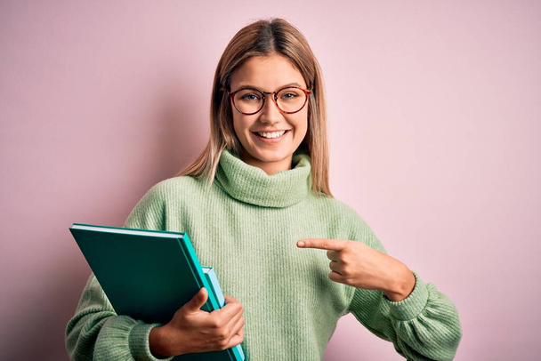 Young beautiful woman wearing glasses holding books over isolated pink background very happy pointing with hand and finger - Valokuva, kuva