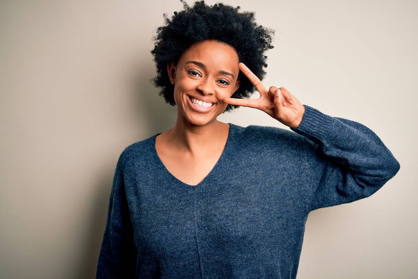 Young beautiful African American afro woman with curly hair wearing casual sweater Doing peace symbol with fingers over face, smiling cheerful showing victory - Foto, Imagem