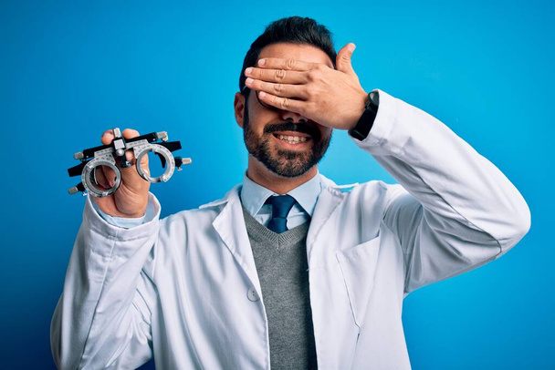 Young handsome optical man with beard holding optometry glasses over blue background smiling and laughing with hand on face covering eyes for surprise. Blind concept. - Photo, Image