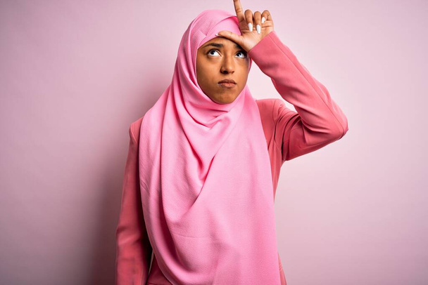 Mladý afroameričan afro žena na sobě muslim hidžáb přes izolované růžové pozadí dělat si legraci z lidí s prsty na čele dělat poražený gesto posměch a urážlivé. - Fotografie, Obrázek