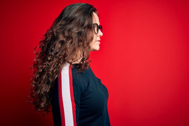 Young beautiful woman with curly hair wearing sweater and glasses over red background looking to side, relax profile pose with natural face with confident smile. - Фото, зображення