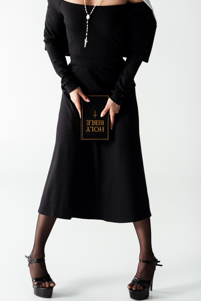 καλλιεργημένη άποψη του σέξι καλόγρια σε μαύρο φόρεμα κρατώντας τη Βίβλο σε γκρι - Φωτογραφία, εικόνα