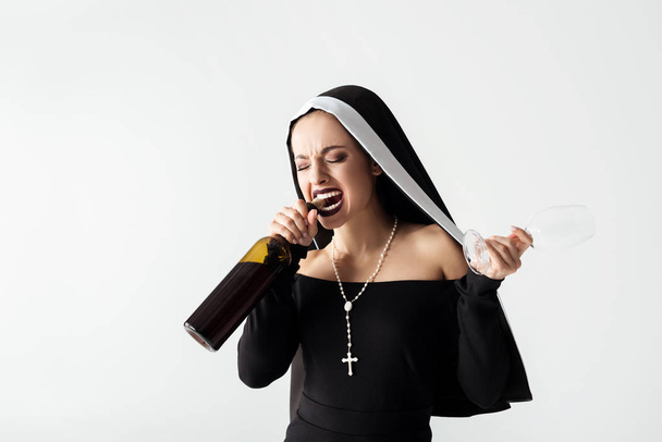 attractive nun holding glass while opening bottle of wine with teeth isolated on grey - Photo, Image